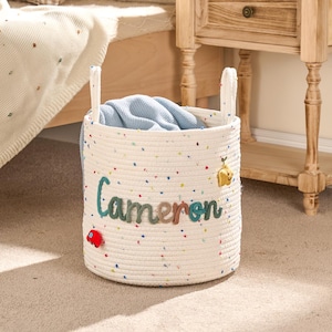 May include: A white rope basket with the name "Cameron" in colourful letters. The basket has two handles and is filled with a blue blanket. A red car and yellow bee are attached to the basket.