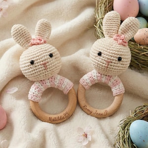 Könnte beinhalten: Zwei handgefertigte Häschen-Rasseln aus Häkelarbeit mit Holzringen, personalisiert mit Namen. Die Häschen sind beige mit rosa Akzenten und einer rosa Blume. Ostereier in Pastellfarben und florale Akzente vervollständigen die Szene.