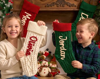 May include: Two cable-knit Christmas stockings featuring a folded cuff and embroidered names. One stocking is cream with the name Olivia in red, and the other is green with a red cuff and the name Jordan in cream. Both have a textured pattern.