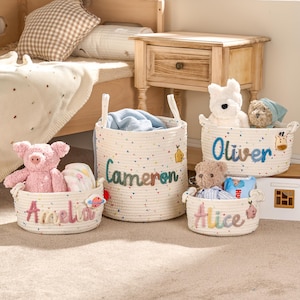 May include: Four cream-coloured woven rope baskets, each with a name embroidered on the front. The baskets contain soft toys and baby items. The names embroidered on the baskets are Amelia, Cameron, Oliver, and Alice.