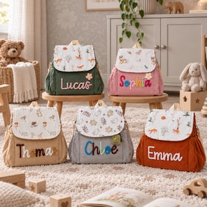 May include: Five corduroy backpacks in green, pink, tan, gray, and orange. Each bag features a white flap with woodland animal prints and a personalized name in colorful, textured lettering. Small decorative accents like flowers or dinosaurs are attached.