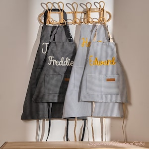 May include: Two personalised aprons hanging on a rattan rack. One is black with white pinstripes and the name "Freddie" embroidered in white. The other is blue and white striped with "Edward" in yellow embroidery. Both have pockets.