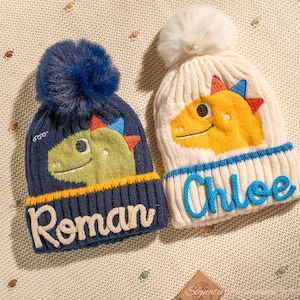 May include: Two knitted dinosaur hats. One is navy blue with a blue pom-pom and the name "Roman" in cream. The other is cream with a white pom-pom and the name "Chloe" in blue. Both have dinosaur faces and are suitable for cold weather.