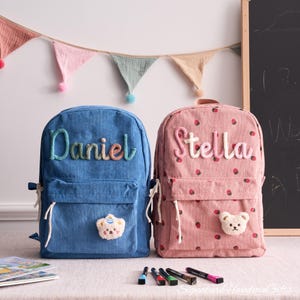 May include: Two corduroy backpacks, one blue with "Daniel" and a bear patch, the other pink with "Stella" and strawberry pattern. Both have front pockets and white drawstrings. A colourful banner hangs in the background.