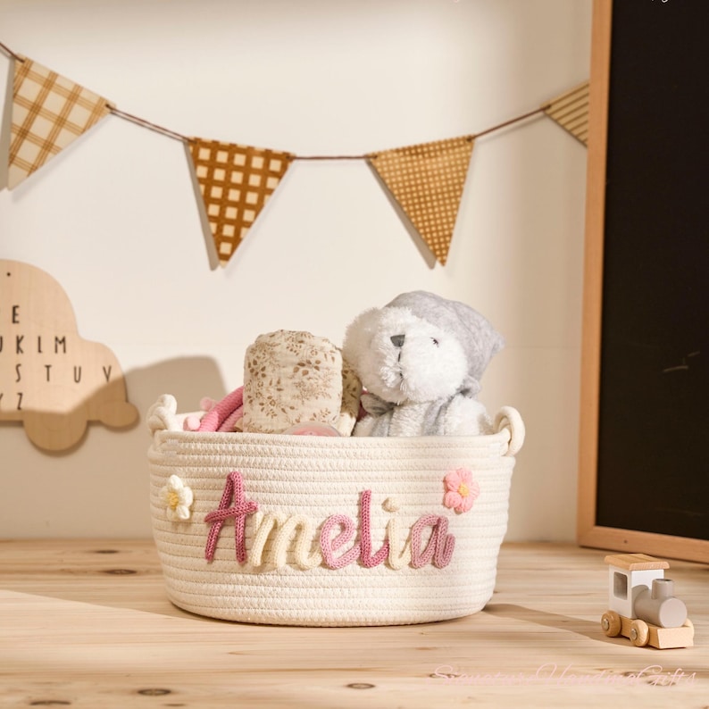 May include: A cream-coloured rope basket, embroidered with the name "Amelia" in pink, and decorated with pink flowers. The basket holds a teddy bear and other soft toys. A wooden train is nearby, with a banner in the background.