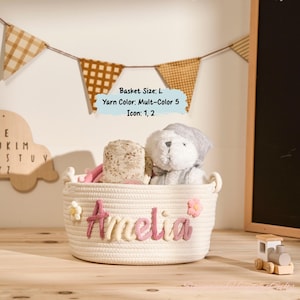May include: A white rope basket with the name "Amelia" in pink and cream-colored letters. The basket contains a stuffed bear and other items. Decorative flowers are attached to the basket. The basket is on a wooden surface.