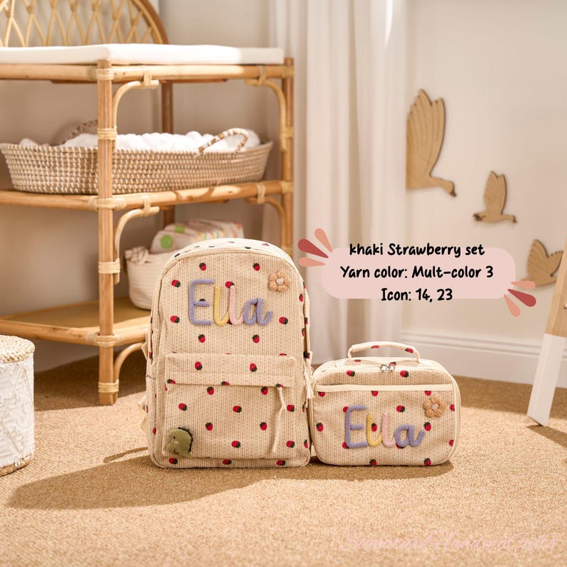 May include: Khaki Strawberry set featuring a backpack and lunch bag. Both items are beige with red strawberry accents and the name "Ella" in pastel colors. The set includes a small flower and a green dinosaur applique.