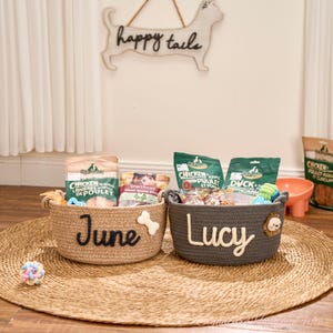May include: Two woven dog treat baskets, one tan and one grey, sit on a round woven mat. The tan basket says "June" and has a bone accent. The grey basket says "Lucy" and has a lion accent. Both baskets contain dog treats and toys.