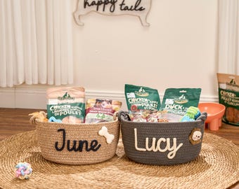 May include: Two woven dog treat baskets, one tan and one gray, sit on a round woven mat. The tan basket says "June" and has a bone accent. The gray basket says "Lucy" and has a lion accent. Both baskets contain dog treats and toys.
