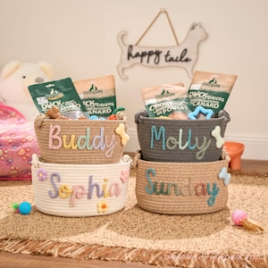 May include: Four woven pet baskets, each with a name embroidered on the front: "Buddy", "Molly", "Sophia", and "Sunday". The baskets are filled with dog treats and toys. The background includes a "happy tails" sign.