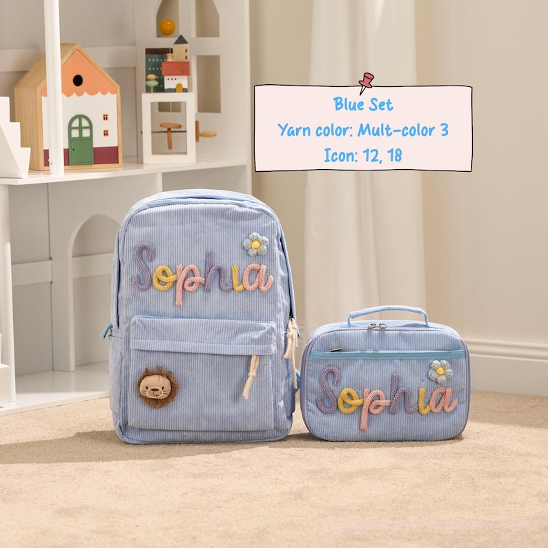 May include: A light blue corduroy backpack and matching lunch bag set. The backpack features the name "Sophia" in colorful embroidery, a flower applique, and a lion patch. The lunch bag also has the name "Sophia" embroidered.