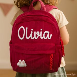 May include: A red corduroy backpack with the name "Olivia" embroidered in white. The backpack has a front pocket, a small decorative patch, and red braided zipper pulls. The bag is designed for children.