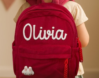May include: A red corduroy backpack with the name "Olivia" embroidered in white. The backpack has a front pocket, a small decorative patch, and red braided zipper pulls. The bag is designed for children.