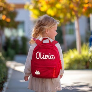 May include: A red corduroy backpack with the name "Olivia" in white script. The backpack has red straps and a small white bunny patch. The child is wearing a sweater and dress.