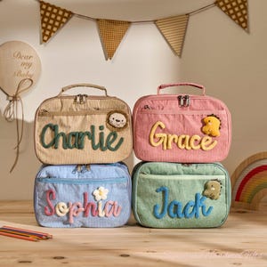 Peut inclure: Quatre sacs à lunch en velours côtelé beige, rose, bleu et vert, chacun avec un nom brodé dans une couleur contrastante. Le sac beige a un appliqué de lion, le sac rose a un dinosaure, le sac bleu a une fleur et le sac vert a un dinosaure.