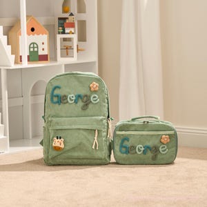 May include: A green corduroy backpack and matching lunch bag set. The backpack features the name "George" in blue and green letters, a giraffe applique, and a flower. The lunch bag also has the name "George" and a flower.