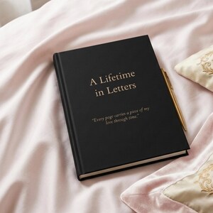 May include: A black journal with the title "A Lifetime in Letters" in gold lettering and a quote. A gold pen lies next to the journal on a pink bedspread. The journal's quote reads, "Every page carries a piece of my love through time."