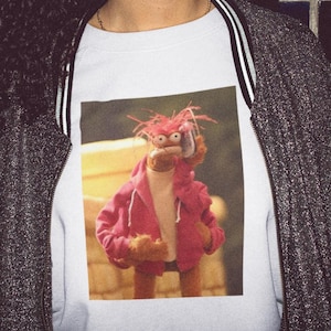 May include: White sweatshirt with a graphic print of a red-haired puppet wearing a pink hoodie and holding a phone to its ear.