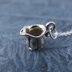 Op de afbeelding: Een zilveren bedel in de vorm van een klein kannetje hangt aan een zilveren ketting. De bedel is gedetailleerd en heeft een gladde, gepolijste afwerking.