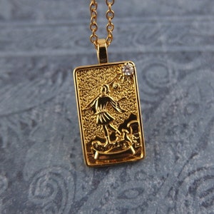 May include: Gold pendant necklace with a rectangular charm featuring a detailed image of a person holding a staff. The charm has a small clear rhinestone at the top.