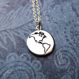 Puede incluir: Un collar de plata con un pequeño colgante redondo que presenta un contorno de la Tierra. El colgante está en una cadena de plata.