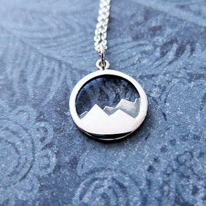 May include: A silver necklace with a round pendant featuring a white silhouette of mountains against a dark gray background.