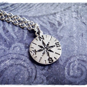 May include: A silver compass necklace with a chain. The compass face has a black star and the letters N, S, E, and W.