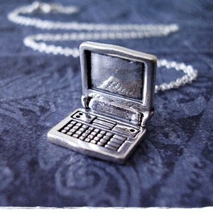 May include: A silver laptop charm necklace. The laptop charm is detailed with a screen, keyboard, and hinge. The chain is silver and has a lobster clasp.
