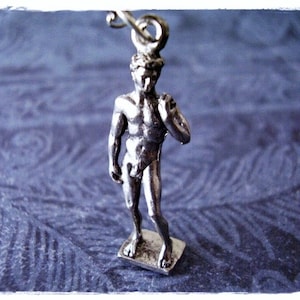 Puede incluir: Encanto de plata de una figura masculina desnuda de pie con una mano en la cadera. La figura es una réplica de la famosa estatua de David de Miguel Ángel.