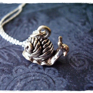 May include: A silver-colored snail pendant necklace. The snail has a detailed shell and is attached to a delicate silver chain. The necklace is displayed on a dark blue textured surface.