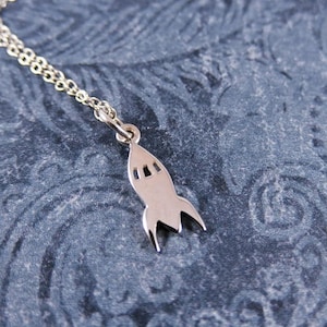 Puede incluir: Un collar de cadena de plata con un colgante de cohete. El colgante es un diseño simple y estilizado con una nariz puntiaguda y aletas.