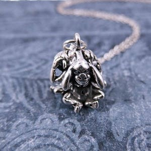 Puede incluir: Collar de plata con un dije de un mono que se tapa los ojos, las orejas y la boca. El mono está sentado sobre una superficie azul texturizada.