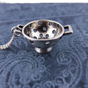 May include: A silver colander charm necklace. The colander is a small, detailed replica of a kitchen colander with holes throughout. The necklace is made of a silver chain.