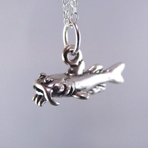 Puede incluir: Collar con colgante de pez plateado. El colgante es una representación detallada de un pez, con aletas, escamas y boca visibles. Cuelga de una fina cadena de plata. Una joya sencilla pero llamativa.