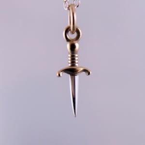 May include: A gold-coloured dagger pendant with a silver blade suspended from a delicate silver chain. The dagger has a detailed hilt and guard, displaying a classic design. The pendant hangs against a soft, neutral background.