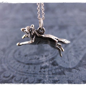 May include: A silver-toned pendant necklace featuring a detailed, running wolf charm. The wolf is depicted mid-stride, suspended from a delicate chain. The pendant is a small, detailed sculpture, showcasing the animal's form. The background is a soft, patterned blue.