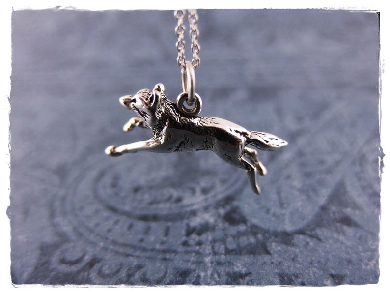 May include: A silver-toned pendant necklace featuring a detailed, running wolf charm. The wolf is depicted mid-stride, suspended from a delicate chain. The pendant is a small, detailed sculpture, showcasing the animal's form. The background is a soft, patterned blue.