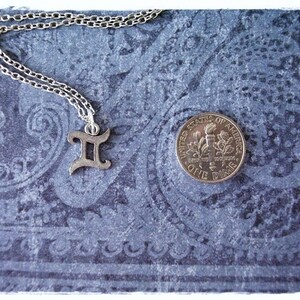 May include: A silver necklace with a Gemini zodiac symbol charm on a blue patterned background. A US dime is also pictured for scale.