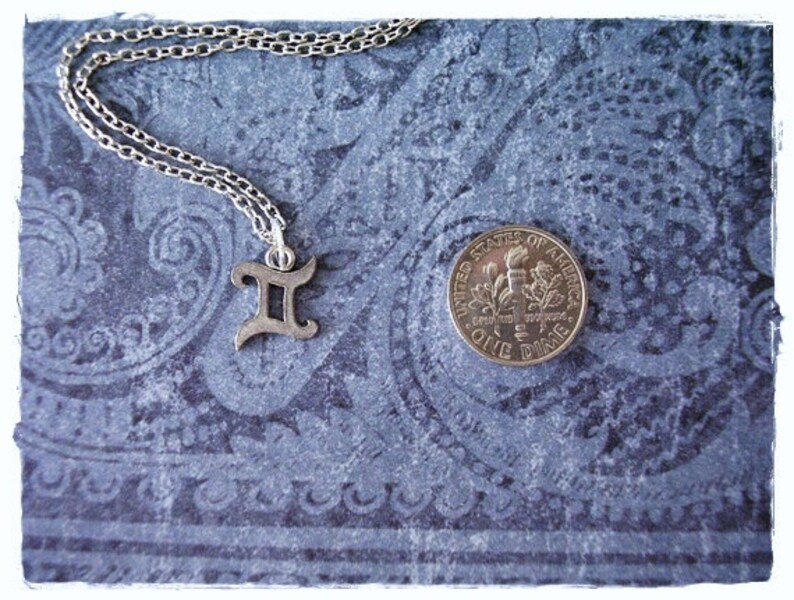 May include: A silver necklace with a Gemini zodiac symbol charm on a blue patterned background. A US dime is also pictured for scale.