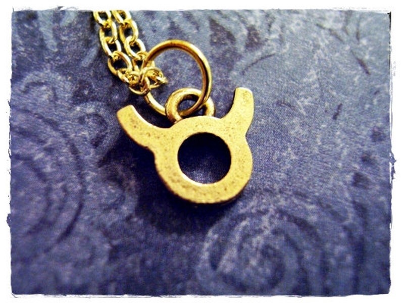 May include: A gold-colored necklace with a Taurus zodiac sign pendant. The pendant is a simple outline of a bull's head.