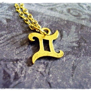May include: Gold-tone necklace with a Gemini zodiac sign pendant. The pendant is a stylized letter 'II' with a decorative flourish.