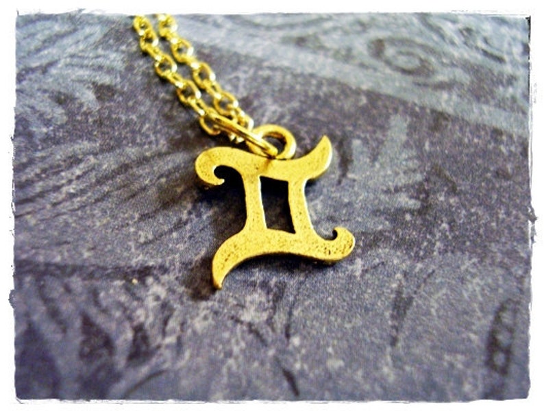 May include: Gold-tone necklace with a Gemini zodiac sign pendant. The pendant is a stylized letter 'II' with a decorative flourish.