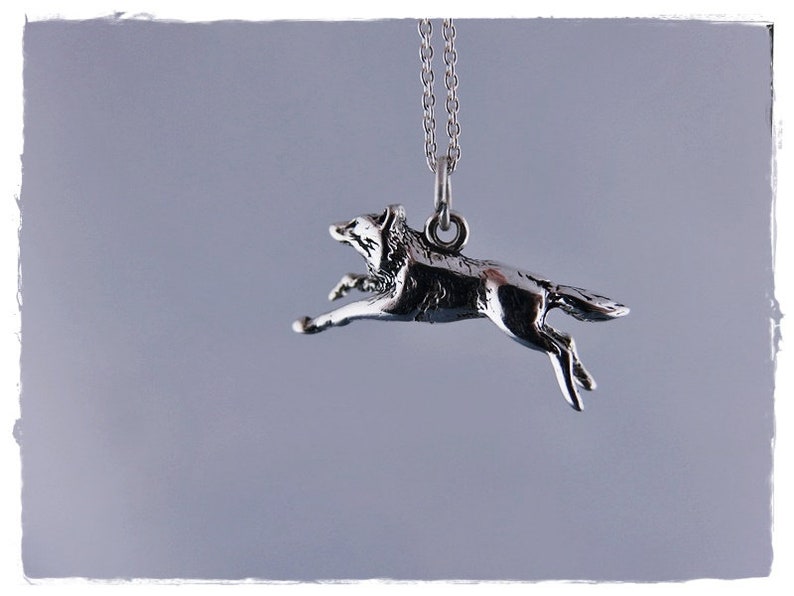 May include: A silver-colored pendant necklace featuring a running wolf charm. The wolf is depicted mid-stride, with its legs extended. The pendant hangs from a delicate silver chain, creating a simple yet striking piece of jewelry.