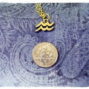 May include: Gold necklace with a wave pendant charm. The pendant is a simple design of three waves. The necklace is on a blue patterned background with a US dime for scale.