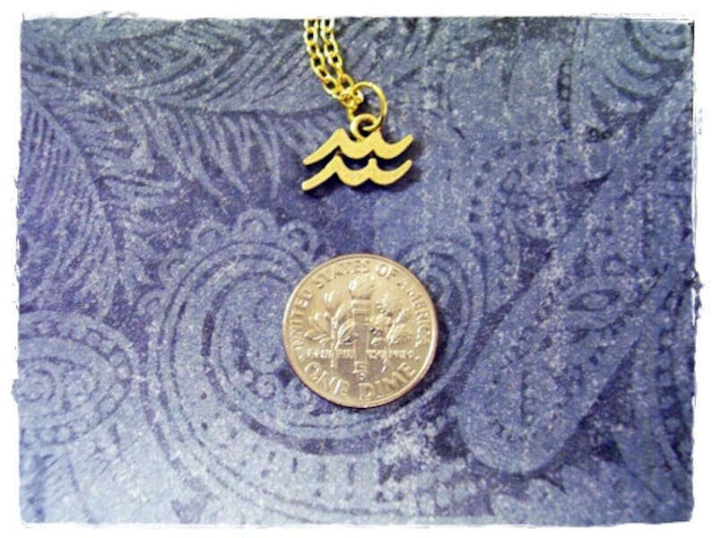 May include: Gold necklace with a wave pendant charm. The pendant is a simple design of three waves. The necklace is on a blue patterned background with a US dime for scale.