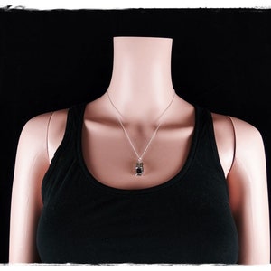 May include: A silver chain necklace with a small, dark gray pendant. The necklace is worn by a mannequin wearing a black tank top.