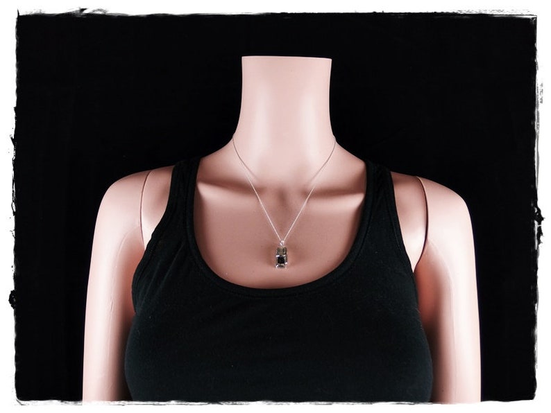 May include: A silver chain necklace with a small, dark gray pendant. The necklace is worn by a mannequin wearing a black tank top.