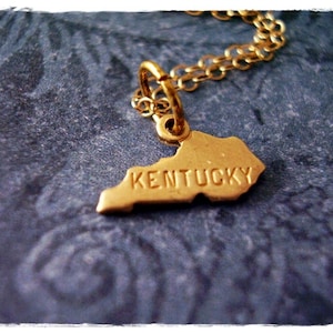 May include: A gold necklace with a pendant shaped like the state of Kentucky. The pendant has the word "Kentucky" engraved on it.