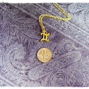 May include: Gold chain necklace with a gold Gemini zodiac sign pendant. The necklace is on a purple patterned background.