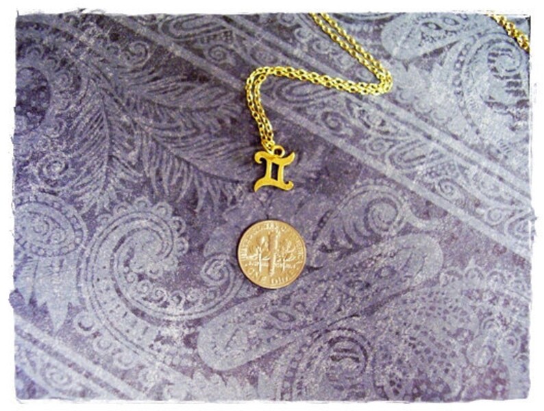 May include: Gold chain necklace with a gold Gemini zodiac sign pendant. The necklace is on a purple patterned background.